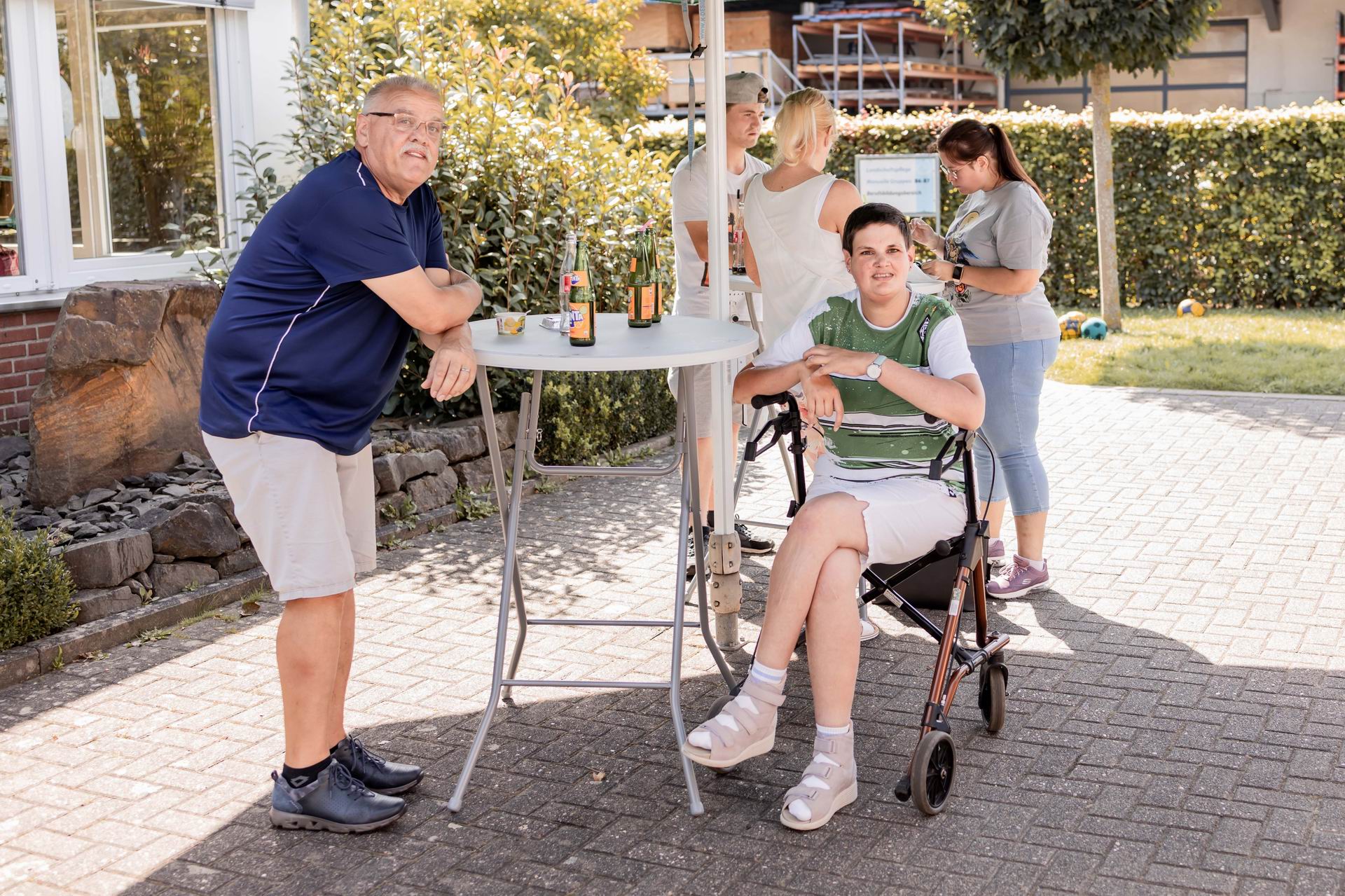 Familie genießt einen sonnigen Tag im Freien, während sie an einem Tisch mit Getränken stehen. Eine Person sitzt im Rollstuhl, umgeben von Angehörigen, die eine entspannte Atmosphäre ausstrahlen.