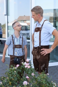 Zwei Männer in traditionellen bayerischen Lederhosen stehen vor einem Gebäude, einer von ihnen lächelt und schaut auf den anderen. Im Vordergrund sind Blumen zu sehen.