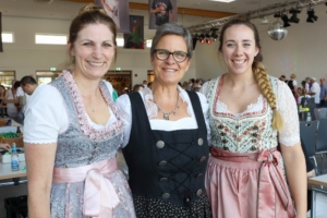 Drei Frauen in traditioneller Tracht lächeln in einem festlichen Ambiente, ideal für kulturelle Veranstaltungen oder Feste.