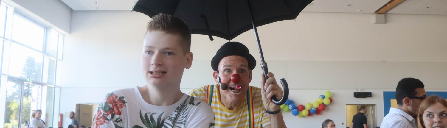 Ein Clown steht mit einem Regenschirm vor einer Menschenmenge. Er hat ein lustiges Outfit an.