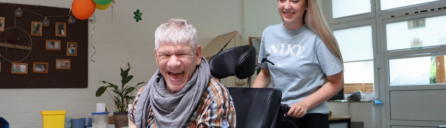 Ein Mann sitzt im Rollstuhl und lacht. Eine Frau steht daneben und lächelt.