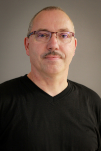 Porträt eines Mannes mit Brille und Schnurrbart, der in einem schwarzen T-Shirt vor einem neutralen Hintergrund steht.