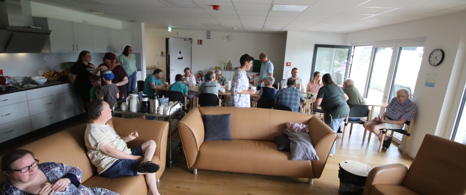 Menschen sitzen in einem Raum. Manche sitzen auf einem Sofa, andere auf Stühlen. Es gibt auch eine Küche im Hintergrund.