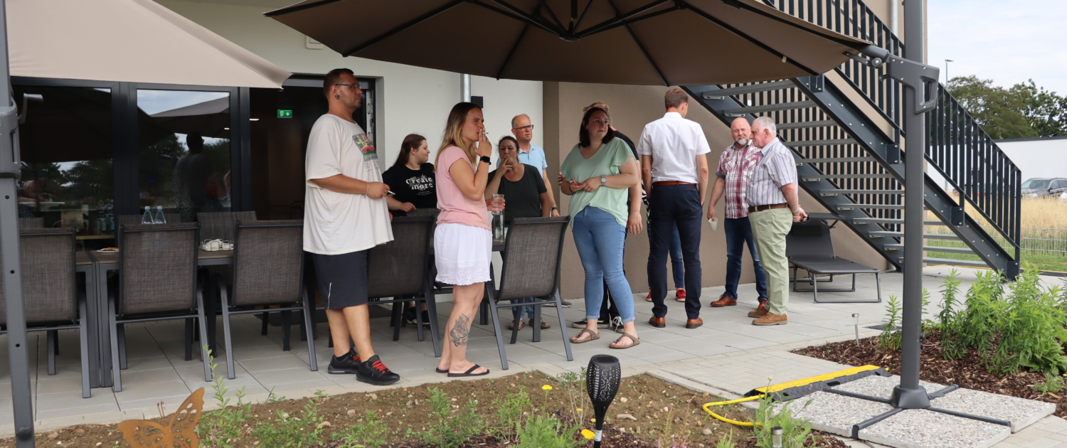 Menschen stehen draußen unter einer überdachten Terrasse. Einige unterhalten sich und lächeln.