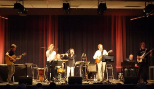 Musikgruppe bei einem Live-Auftritt auf der Bühne, bestehend aus mehreren Musikern, die verschiedene Instrumente spielen. Im Hintergrund sind Instrumente und ein Mikrofon zu sehen. Die Bühnenbeleuchtung schafft eine warme Atmosphäre.