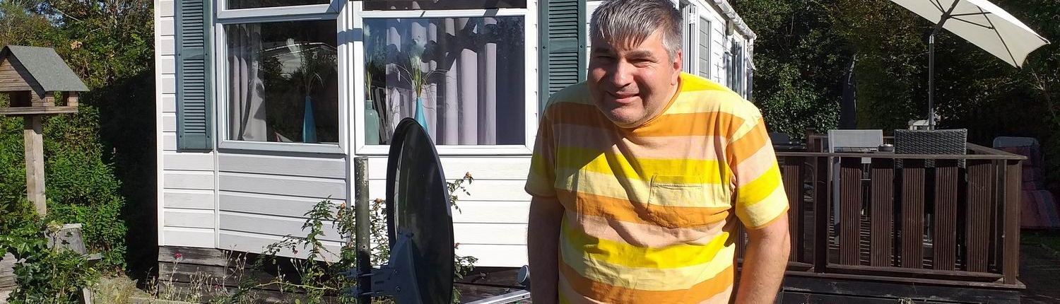 Ein Mann steht vor einem Häuschen. Er trägt ein gelbes T-Shirt und eine Hose mit Blumen. Hinter ihm ist ein Garten zu sehen.