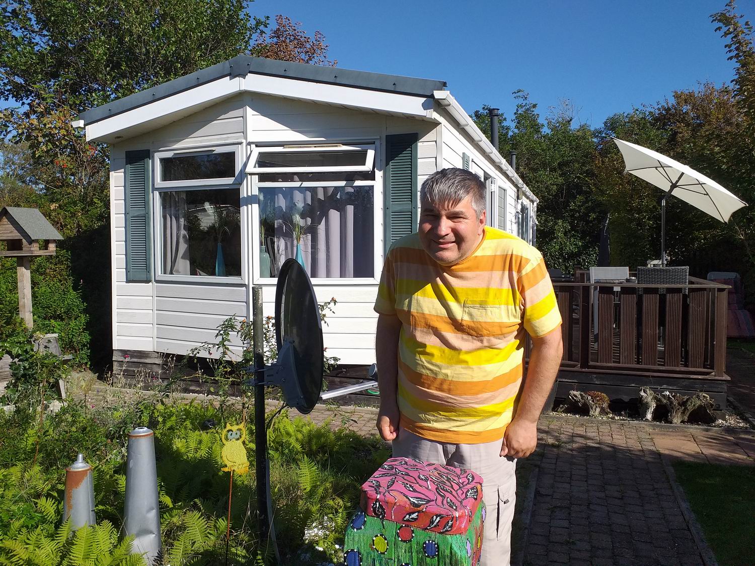 Ein Mann steht vor einem Häuschen. Er trägt ein gelbes T-Shirt und eine Hose mit Blumen. Hinter ihm ist ein Garten zu sehen.