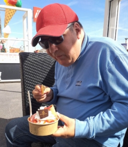 Älterer Mann mit roter Kappe sitzt im Freien und genießt eine Schüssel mit Essen.