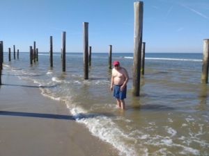 Ein Mann steht im Wasser am Strand. Er trägt eine rote Mütze und eine kurze Hose. Hinter ihm sind Holzpfähle im Wasser zu sehen.