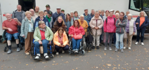 Gruppe von Menschen unterschiedlichen Alters, darunter Senioren in Rollstühlen und Stehenden, die gemeinsam im Freien posieren. Sie tragen bunte Kleidung und lächeln, was eine freundliche Gemeinschaftsatmosphäre vermittelt.