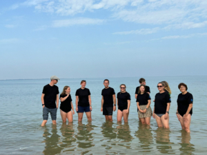 Eine Gruppe von Leuten steht im flachen Wasser am Strand. Der Himmel ist blau.