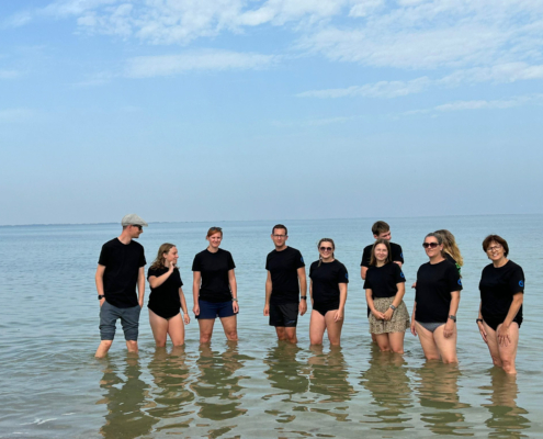 Eine Gruppe von Leuten steht im flachen Wasser am Strand. Der Himmel ist blau.