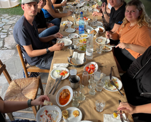 Eine Gruppe von Personen sitzt an einem Tisch im Freien und genießt ein reichhaltiges Essen mit verschiedenen Gerichten und Getränken.