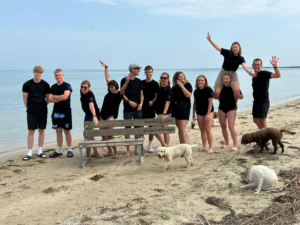 Eine Gruppe von Jugendlichen steht auf einem Strand. Ein Hund steht neben ihnen.