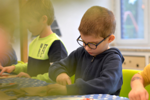 Ein Junge mit Brille arbeitet konzentriert an einer Bastelaktivität in einem Klassenzimmer. Im Hintergrund ist ein weiterer Schüler zu sehen.