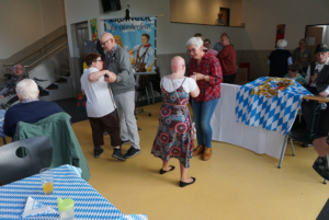 Gruppenszene bei einer Feier in einem Gemeinschaftsraum, in der Menschen lachen und tanzen. Bunte Tische mit Dekorationen sind im Hintergrund sichtbar, was eine festliche Atmosphäre schafft.