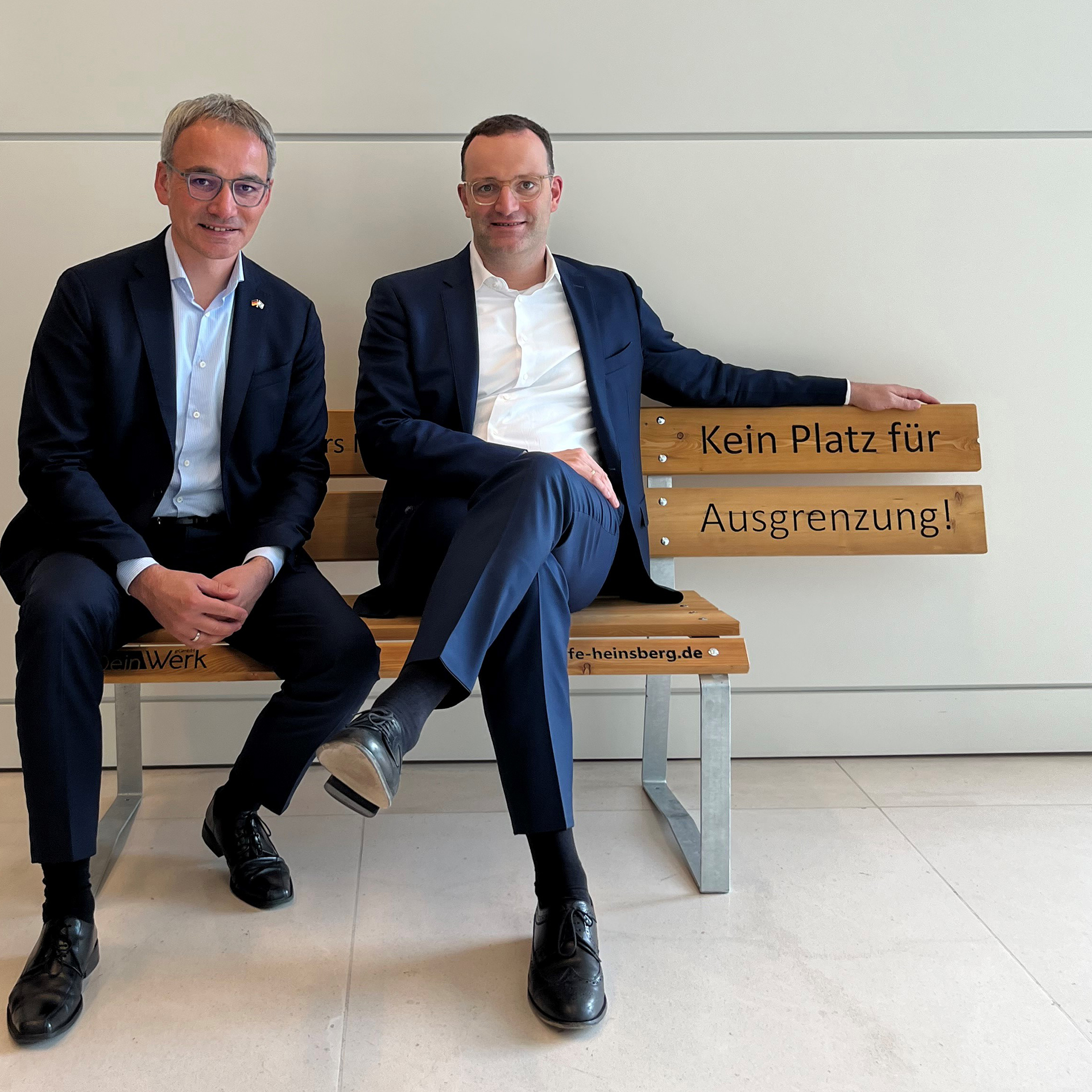 Zwei Geschäftsleute in Anzügen sitzen auf einer Holzbank in einem modernen Büro. Sie lächeln und wirken entspannt, während sie sich unterhalten.