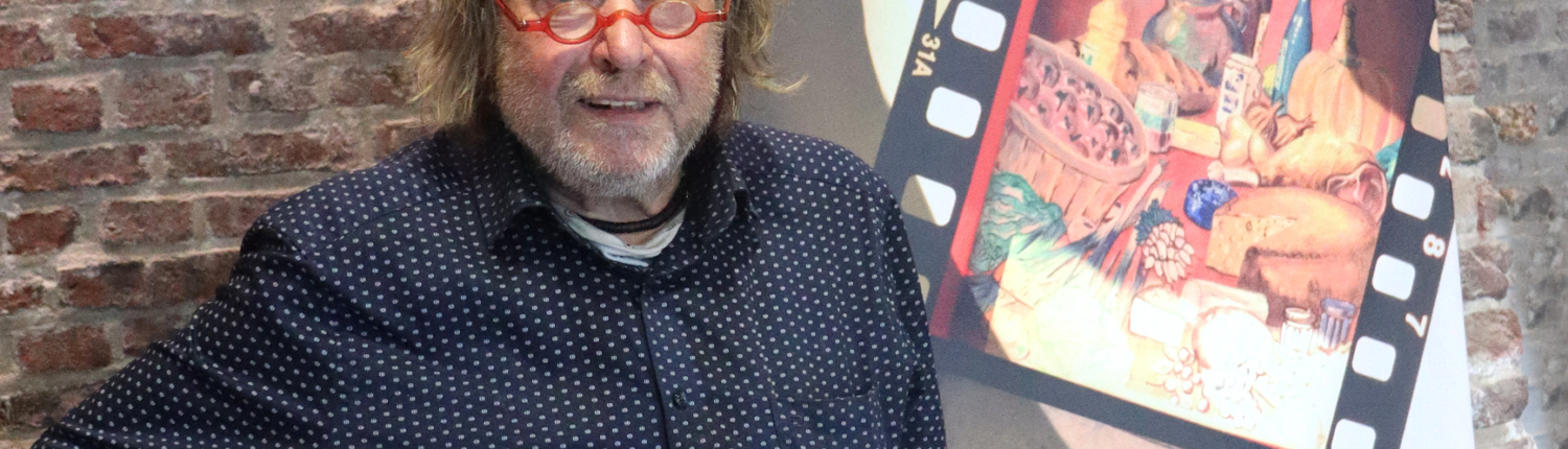 Ein Mann steht vor einer Mauer. Er hat eine Brille und graues Haar. Er trägt ein blaues Hemd und eine braune Hose.