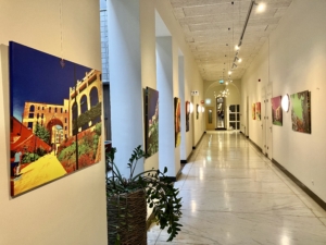 Ein heller Flur mit Kunstwerken an den Wänden, der in einer Galerie oder einem Museum zu sehen ist. Pflanzen stehen am Boden, während das Licht durch große Fenster strömt und die Farben der Gemälde betont.