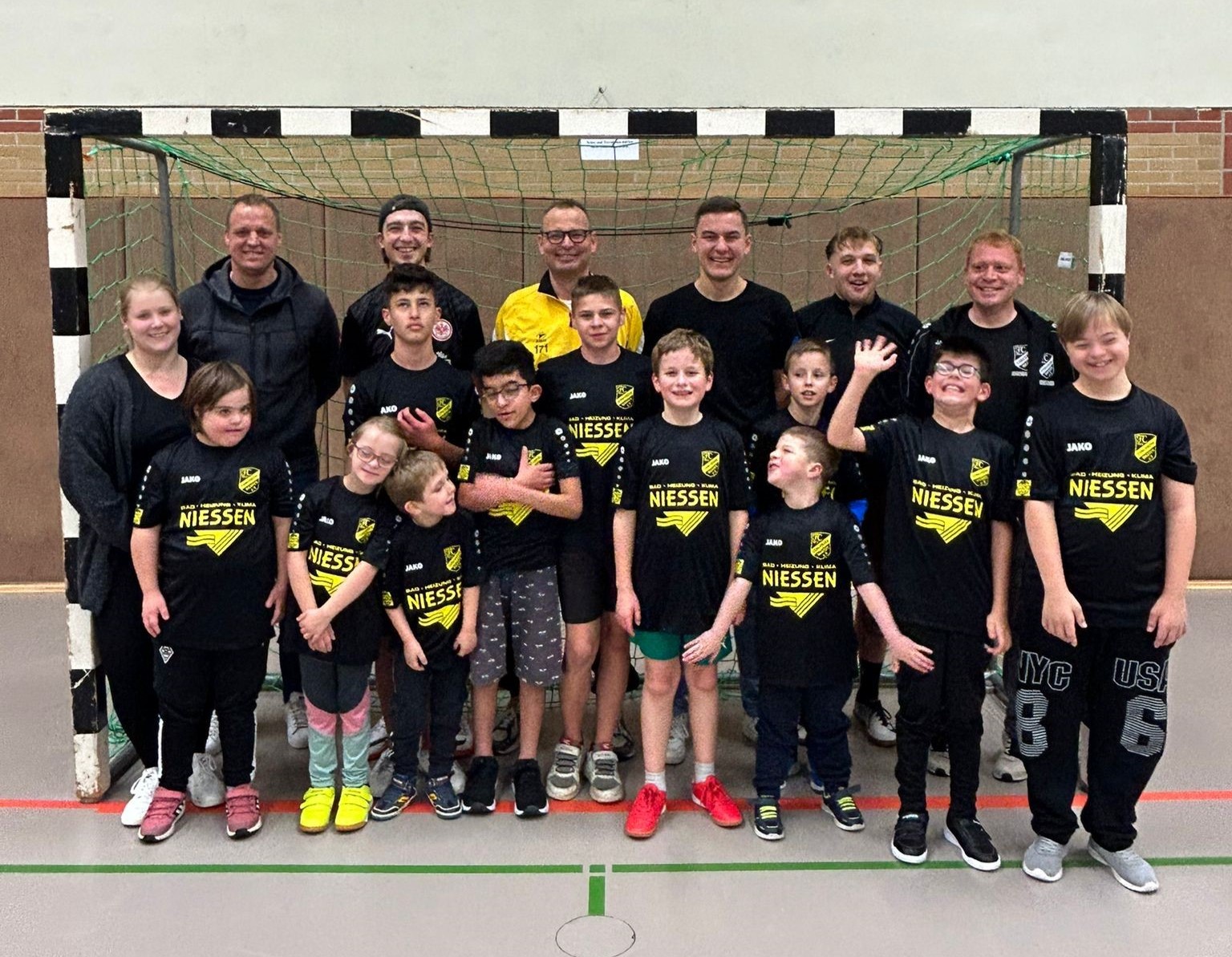 Eine Jugendfußballmannschaft steht vor einem Tor. Die Kinder tragen schwarze Trikots und gelbe Hosen. Ein Mann steht mit ihnen.