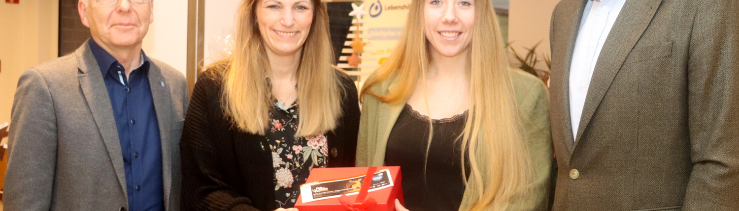 Vier Personen stehen in einem festlichen Raum und präsentieren ein Geschenk. Zwei Frauen halten eine rote Geschenkbox, während die anderen beiden Männer lächeln und sie unterstützen. Die Szene deutet auf eine Feier oder besondere Veranstaltung hin.