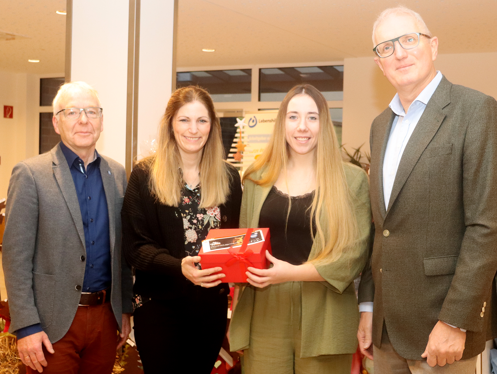 Vier Personen stehen in einem festlichen Raum und präsentieren ein Geschenk. Zwei Frauen halten eine rote Geschenkbox, während die anderen beiden Männer lächeln und sie unterstützen. Die Szene deutet auf eine Feier oder besondere Veranstaltung hin.