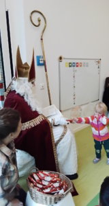 Ein Kind interagiert mit einer Person, die als Nikolaus verkleidet ist, während eine weitere Person im Hintergrund zuschaut. Die Szene spielt in einem hellen Raum mit einer Tafel im Hintergrund.