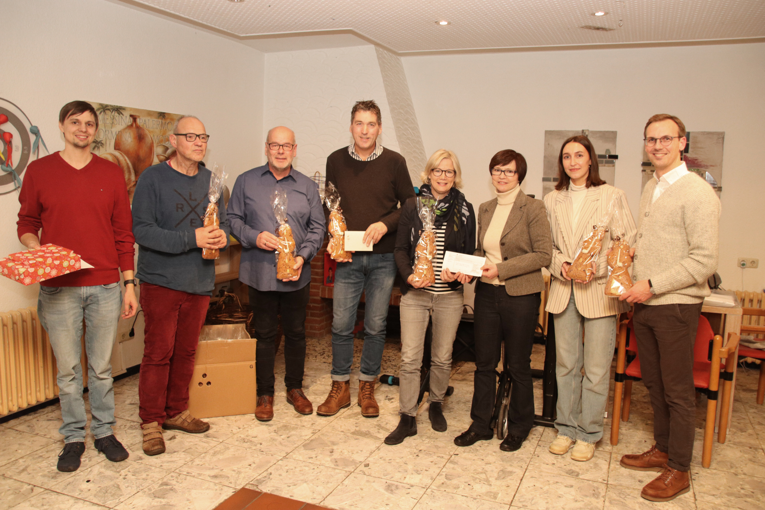 Eine Gruppe von Menschen steht in einem Raum. Einige halten Päckchen oder Umschläge in der Hand.
