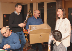Familie feiert zusammen, während ein Mann mit einer großen Kiste in der Mitte des Raumes steht. Im Hintergrund sind weitere Personen zu sehen, die an der Feier teilnehmen. Die Atmosphäre ist festlich und gesellig.