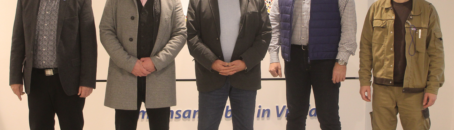 Gruppenfoto von fünf Männern in formeller Kleidung, die an einer Veranstaltung teilnehmen. Im Hintergrund ist ein dekoratives Wandmuster sichtbar.