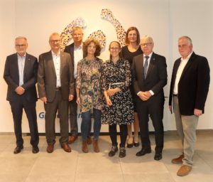 Gruppenfoto von sieben Personen in formeller Kleidung, die in einem modernen Raum posieren, mit einer künstlerischen Wandgestaltung im Hintergrund.