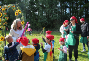 Kinder mit roten Mützen und bunten Overalls spielen im Freien, während eine Betreuerin mit einem langen Ballon eine Aktivität anleitet. Im Hintergrund sind Sonnenblumen und andere Kinder zu sehen.
