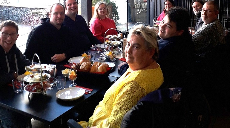 Gruppe von Menschen, die an einem Tisch in einem Restaurant sitzen und gemeinsam essen. Die Atmosphäre ist freundlich und gesellig, mit Blick auf eine Fensterfront.