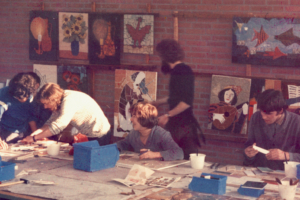 Künstler arbeiten in einem Atelier an verschiedenen Kunstprojekten, umgeben von Gemälden an den Wänden und Materialien auf dem Tisch.