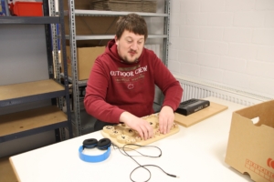 Ein Mann schneidet mit einem Messer etwas aus. Er trägt ein rotes T-Shirt. Hinter ihm stehen Kartons.