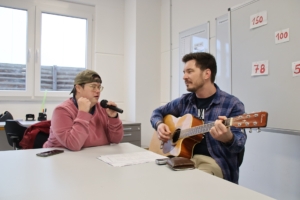 Zwei Personen sitzen an einem Tisch. Eine Person spielt Gitarre, die andere hält ein Mikrofon.