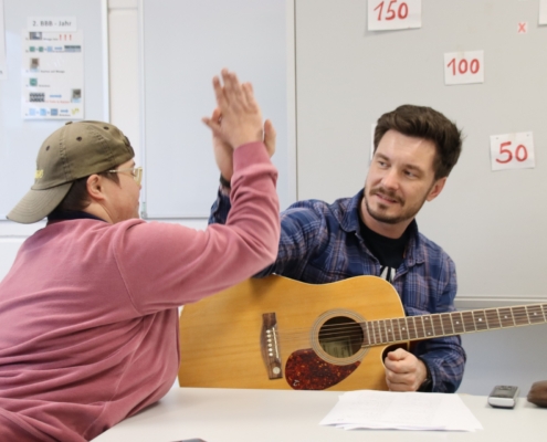 2023-12-15_Mike Musikgruppe (68) Zwei Personen in einem Raum, einer hochfährt die Hand zur Begrüßung, während der andere auf einer Gitarre spielt. Beide zeigen Freude und Interaktion, was eine positive Lern- oder Therapiesituation andeutet.