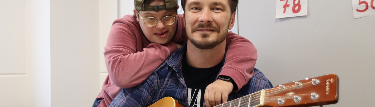 Zwei Männer posieren in einem Raum, einer spielt Gitarre, während der andere ihn umarmt. Beide lächeln, was eine freundliche und entspannte Atmosphäre vermittelt.