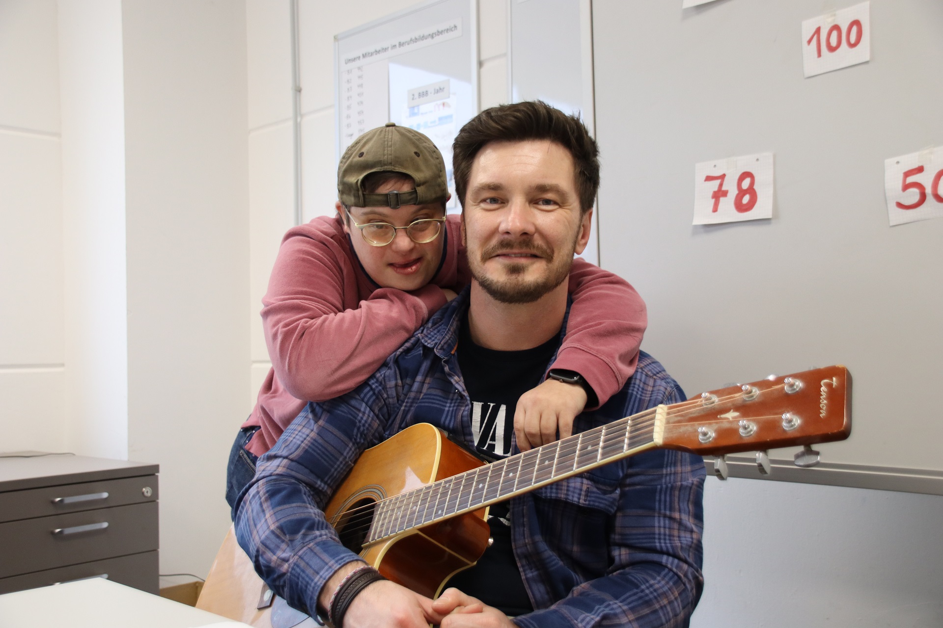 Zwei Männer posieren in einem Raum, einer spielt Gitarre, während der andere ihn umarmt. Beide lächeln, was eine freundliche und entspannte Atmosphäre vermittelt.