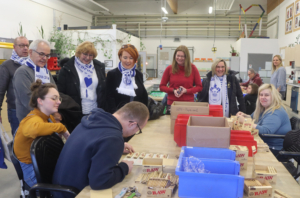 Menschen stehen an Tischen mit vielen Kartons und Packungen. Es sieht aus wie eine Ausgabestelle.