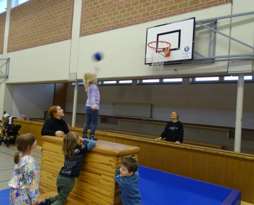 Kinder turnen im Hallen. Ein Mädchen klettert auf eine Bank. Eine Frau schaut zu.