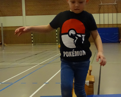 Ein Kind in einem Pokémon-T-Shirt balanciert auf einer Turnbank in einer Sporthalle.
