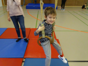 Ein Junge schwingt an Turnringen in einer Sporthalle, während eine weitere Person im Hintergrund steht. Die Matte auf dem Boden ist rot und blau, was eine sichere Umgebung für das Spielen und Trainieren bietet.