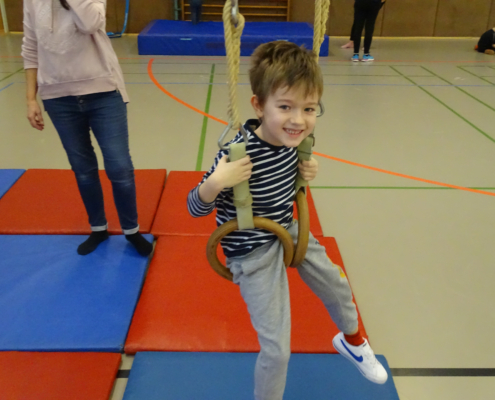 Ein Junge schwingt an Turnringen in einer Sporthalle, während eine weitere Person im Hintergrund steht. Die Matte auf dem Boden ist rot und blau, was eine sichere Umgebung für das Spielen und Trainieren bietet.