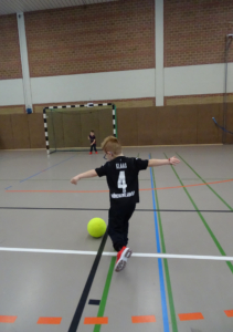 Ein Junge wirft einen Ball in einer Sporthalle. Er trägt ein schwarzes T-Shirt und eine kurze Hose.