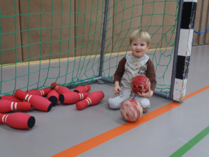 Ein Kind sitzt auf dem Boden und hält zwei Bälle. Hinter dem Kind sind noch mehr Bälle zu sehen.