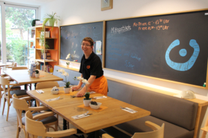 Eine Person steht in einem Restaurant. Es gibt Tische und Stühle. Die Person trägt eine schwarze Weste.