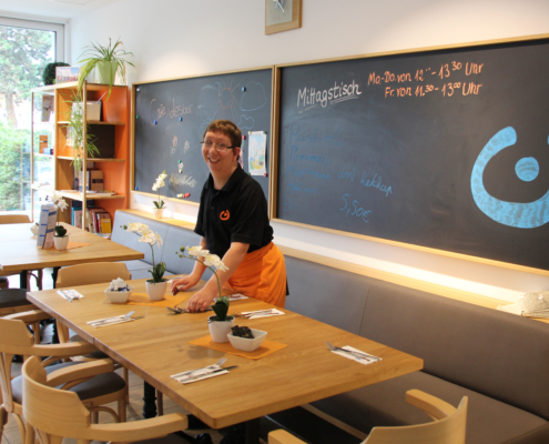 Eine Person steht in einem Restaurant. Es gibt Tische und Stühle. Die Person trägt eine schwarze Weste.