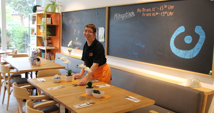Cafe Lesbar eröffnet Eine Person steht in einem Restaurant. Es gibt Tische und Stühle. Die Person trägt eine schwarze Weste.