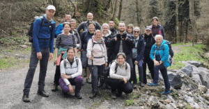 Gruppe von Wanderern auf einem Wanderweg in der Natur, umgeben von Bäumen, lächelnd und posierend für ein Gruppenfoto.